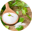 White granulated sugar in a wooden spoon with a small green leaf, surrounded by fresh green leaves and burlap fabric.