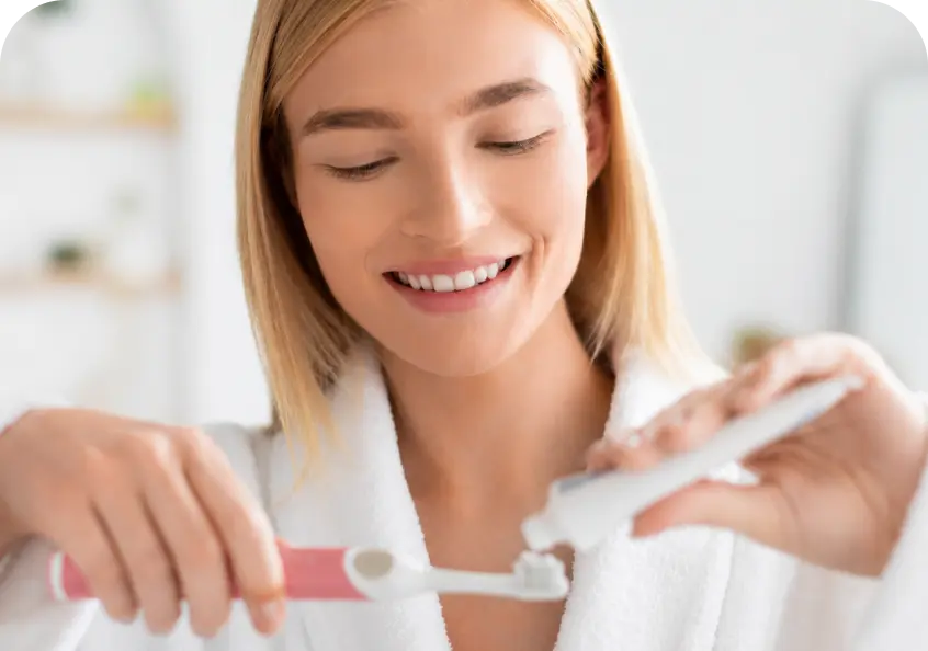 A smiling blonde woman in a white bathrobe applies toothpaste from a white tube onto a pink-handled toothbrush in a bright bathroom setting.