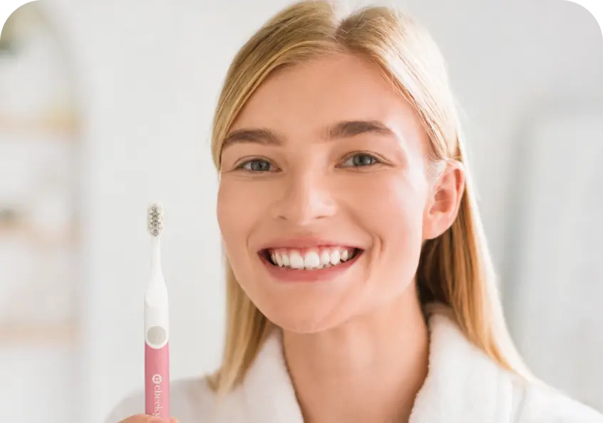 A smiling blonde woman in a white robe holds up a pink and white electric toothbrush with a visible Sonicwave logo, set against a bright, softly blurred bathroom background.